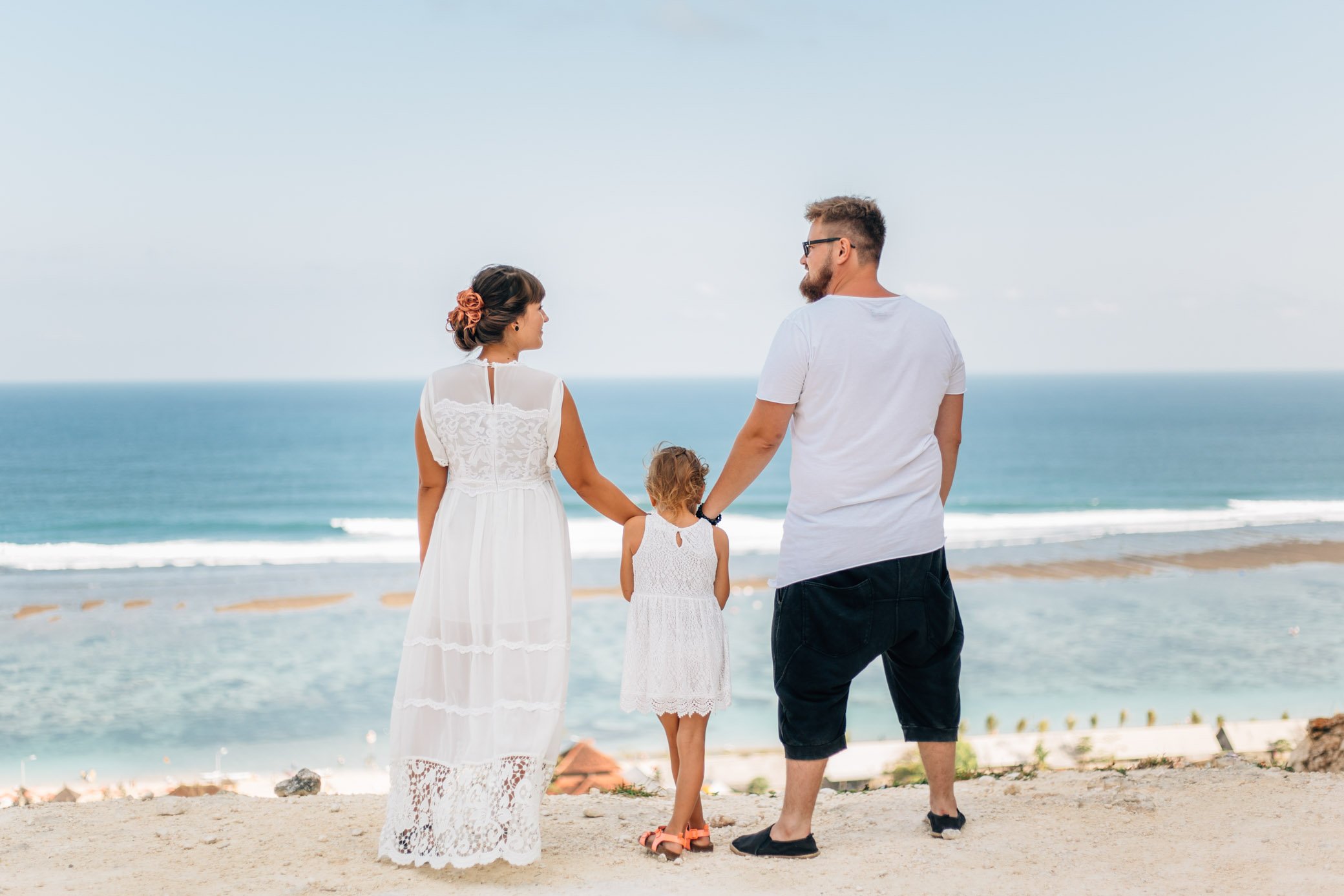 
Mom, Dad and daughter stand with their backs to the camera, they look at the blue ocean. Photosession was held on the Balinese beach Pandava