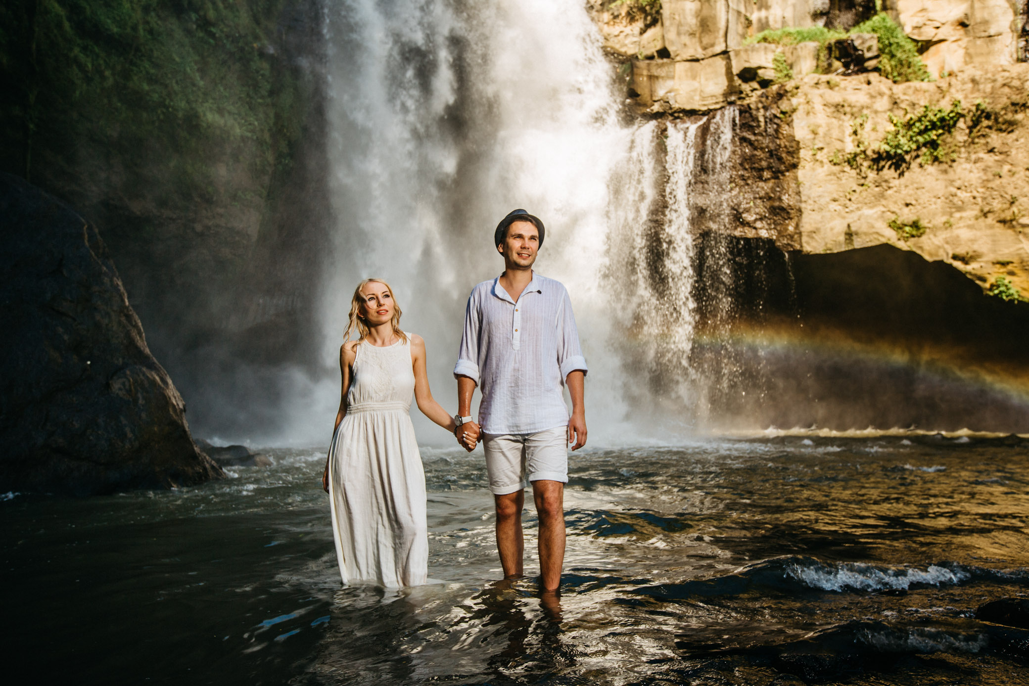 
Photosession at the Tegenungan waterfall, Bali island. The guys are holding hands, watching the distance. A guy in a hat, a girl in a white summer dress with wet hair