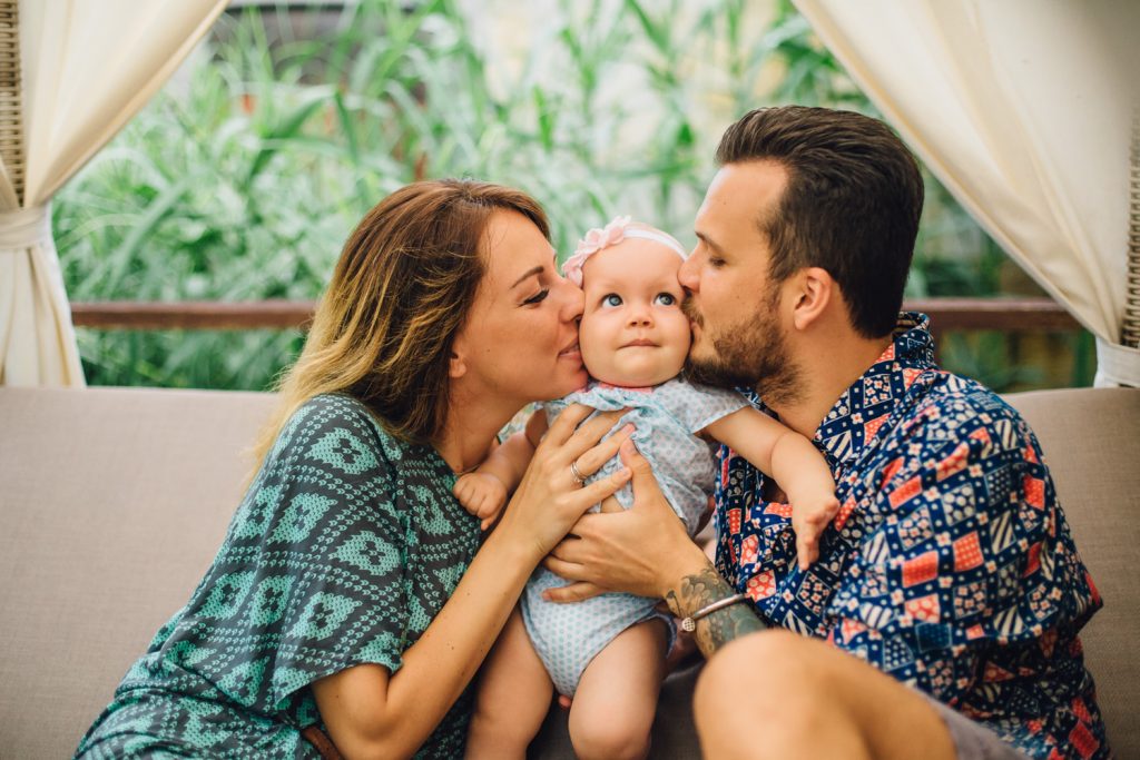 
Mom in a green dress kisses the baby on the cheek. Dad in a bright shirt with a beard holds the baby in his arms. Happy family photo session with a small child in the villa