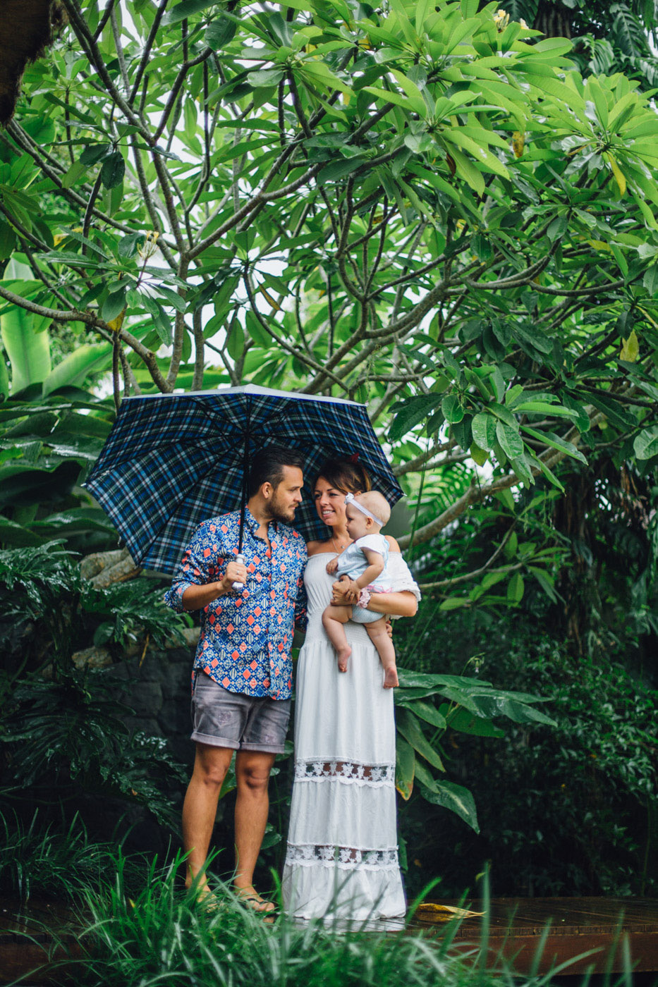 Фотосессия с маленьким ребенком в дождь. Мама, папа и ребенок стоят под синим клетчатым зонтом. Вокруг джунгли и зелень. Тропический дождь